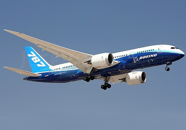 A blue and white Boeing 787 Dreamliner airplane flying against a clear blue sky.
